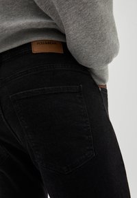 Pantalones de mezclilla negros con una textura suave, que cuentan con dos bolsillos traseros y una etiqueta de cuero marrón claro con "PULL&BEAR." Acompañados de un sudadera gris.