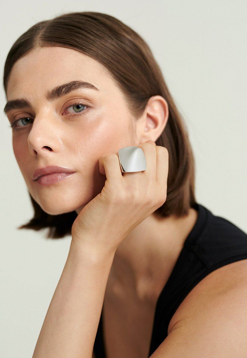 Femme aux cheveux bruns courts reposant le menton sur le poing, portant une grande bague carrée argentée brillante et un haut noir sans manches.