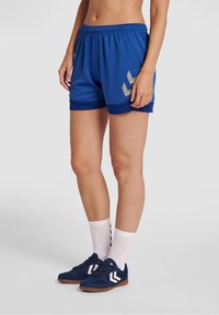 Blå atletiska shorts med mesh-textur och en diagonalt grå logotyp. Kombineras med marinblå sneakers och vita ankelsockor med svarta ränder.