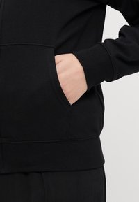 Črna bombažna zip-up kapuca z rebrastimi manšetami in pasom, ki ima stransko žepe spredaj, gladko teksturo in minimalističen dizajn.