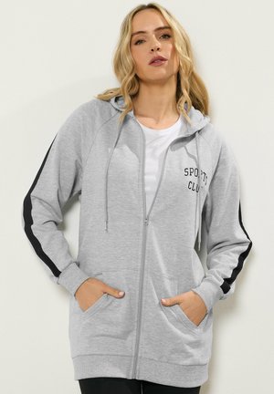 Femme portant un sweat à capuche zippé gris clair avec des bandes noires sur les manches, le texte "SPORTS CLUB", les mains dans les poches, et un T-shirt blanc en dessous.