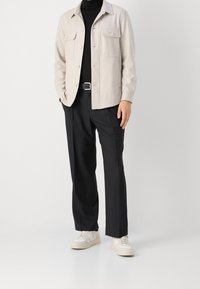 Chaqueta abotonada color beige claro con dos bolsillos en el pecho, combinada con pantalones negros y zapatillas color crema con acentos beige.