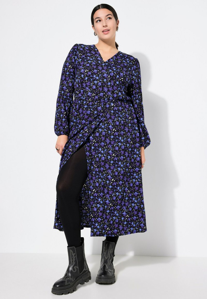 Abito con stampa floreale in viola e blu, con maniche lunghe, scollatura a V e uno spacco laterale, abbinato a leggings neri e stivali neri chunky.
