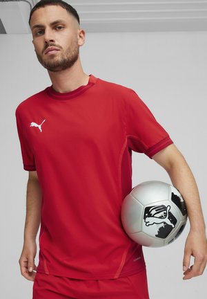 Rode sportieve t-shirt met een ronde hals, korte mouwen en Puma-logo; gladde stof. Speler houdt een zilveren voetbal met zwarte accenten vast.