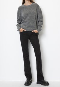 Maglione grigio a maglia con orlo a coste, abbinato a jeans neri a gamba dritta. Scarpe nere con suola spessa completano l'outfit. Sfondo bianco semplice.