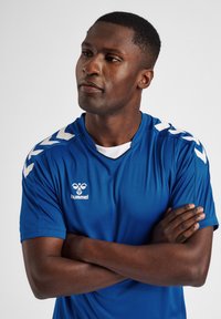 Blå sportsjersey i let stof med hvidt Hummel-logo og chevron-detaljer på ærmerne, samt en runde halsudskæring.