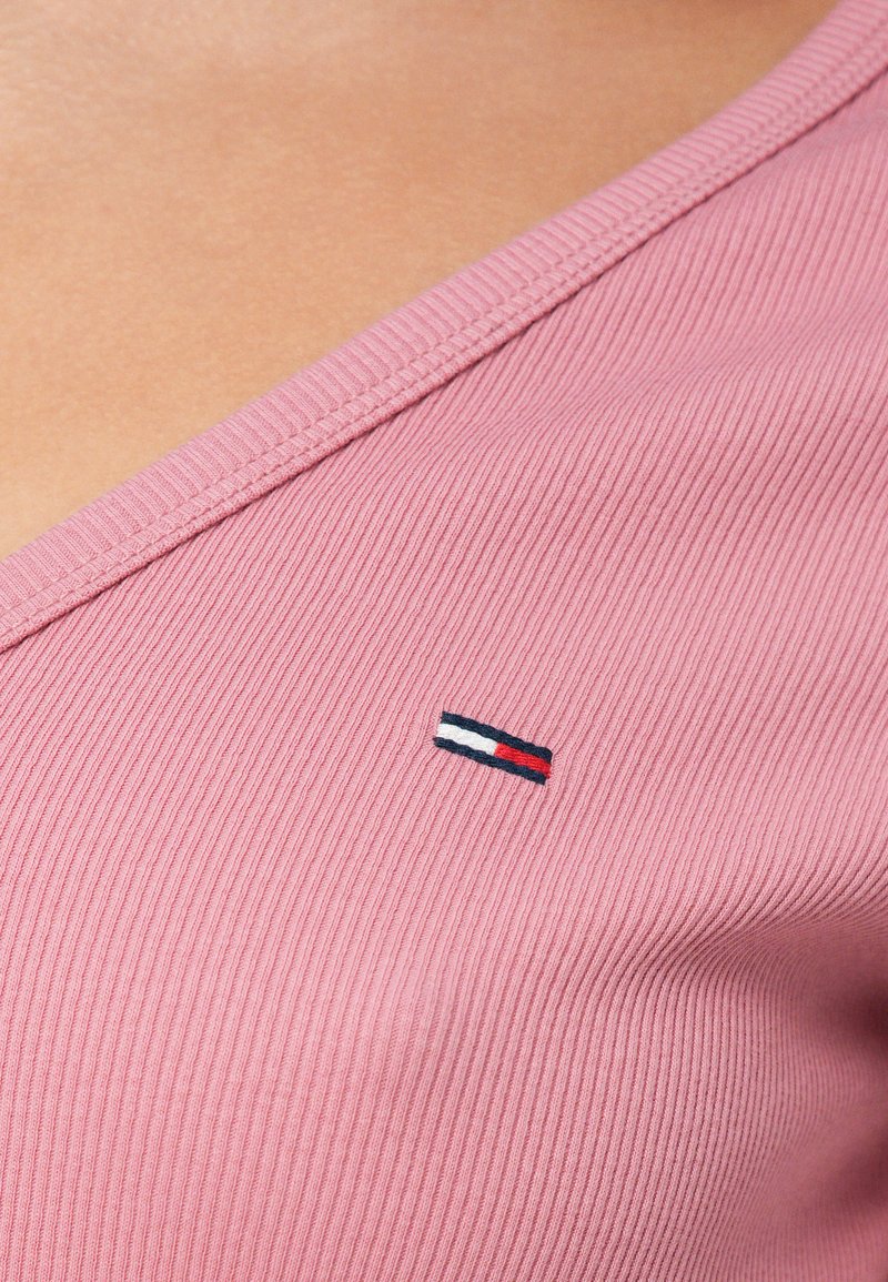 Rosa, geripptes V-Ausschnitt-Top mit kleinem, auf der linken Seite eingesticktem Logo in Marineblau, Weiß und Rot. Glatte Textur.