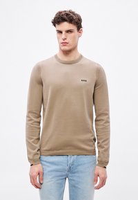 Beżowy sweter z okrągłym dekoltem, ściągaczami na rękawach i u dołu. Na piersi znajduje się małe czarne logo "BOSS". Noszony z jasnoniebieskimi dżinsami.