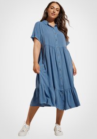 Abito midi blu con bottoni, maniche corte, vita arricciata e gonna a strati. Realizzato in tessuto leggero. Indossato con sneaker bianche.