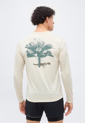 Homme portant une chemise beige à manches longues avec un graphisme d'une grande plante verte feuillue avec des yeux et un petit lézard disant "CHILL" dans le dos.