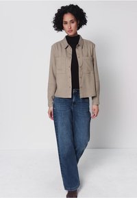 Frau mit lockigem Haar, die vorwärts geht und eine beige knöpfbare Hemdjacket, ein schwarzes Top, weit geschnittene blaue Jeans und braune Schuhe trägt.