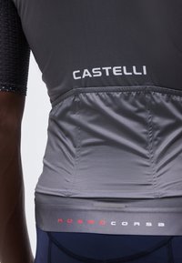 Közeli kép egy személyről, aki fekete és szürke Castelli kerékpáros mezben van, amelyen az alsó hátsó részen a "ROSSOCORSA" felirat olvasható.