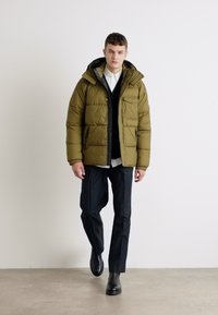 Oliwkowa kurtka puchowa z kapturem, o pikowanej fakturze i zamku błyskawicznym z przodu. W zestawie z czarnymi spodniami i prostymi czarnymi botkami.