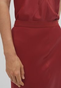 Bordeauxfarbenes Kleid mit einer glatten Satinoberfläche, einer taillierten Silhouette mit einem strukturierten Band und einem minimalistischen Design. Nahaufnahme einer Hand auf dem Stoff.