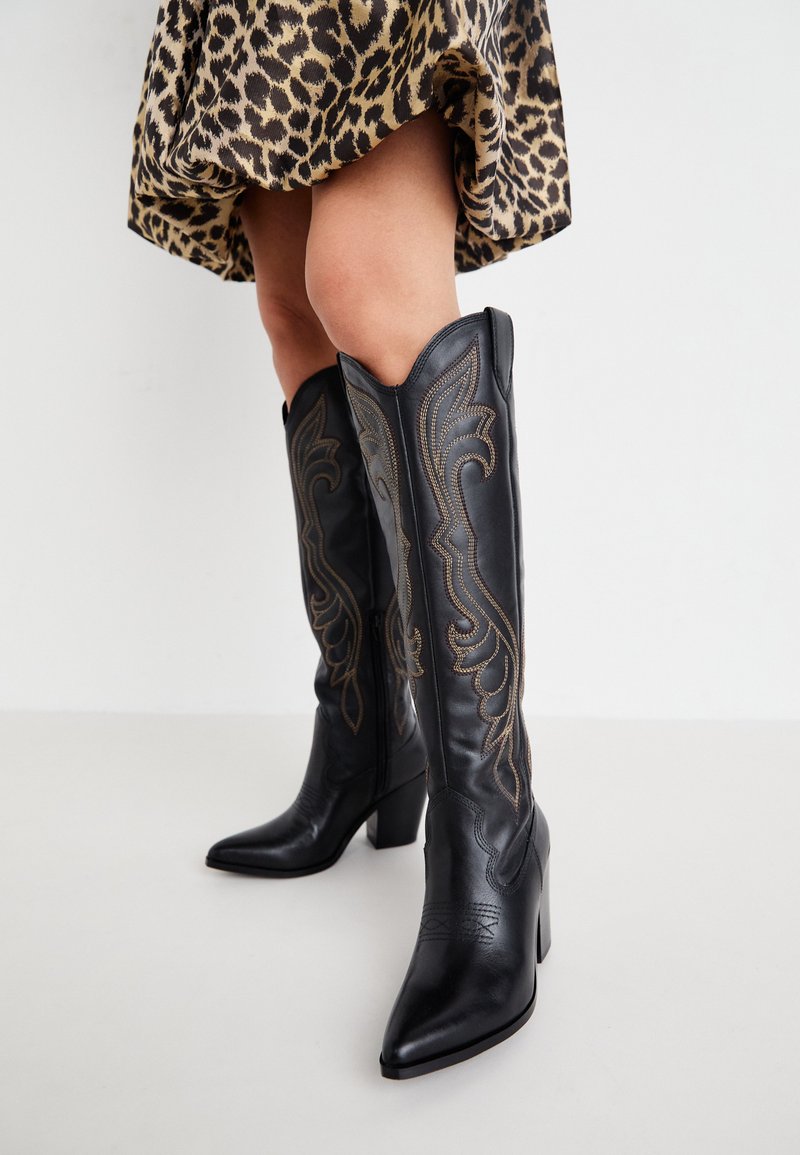 Gambe che indossano stivali da cowboy neri in pelle al ginocchio con cuciture decorative, abbinati a una mini gonna in stampa leopardata su uno sfondo bianco.