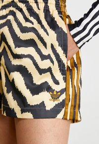 Mönstrade shorts i beige, svart och brunt med en vågig design; har en broderad guld Adidas-logga och kontrasterande sidostreck.
