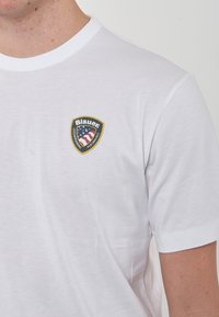 T-shirt bianco in cotone con collo rotondo, caratterizzato da un logo scudetto ricamato con accenti blu, rossi e bianchi sulla parte sinistra del petto.