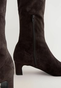 Bottes en suede marron jusqu'au genou avec une fermeture éclair sur le côté, présentant une surface texturée et un talon bas et épais pour la stabilité.