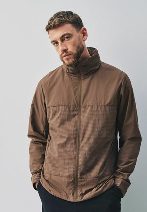 Man with short brown hair and beard wearing a brown zip-up jacket with high collar, standing with hands in pockets against a plain background.