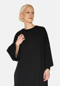 Schwarzes, oversized Kleid aus weichem Stoff, mit weiten Ärmeln, rundem Halsausschnitt und glatter Textur. Einfaches, modernes Design.