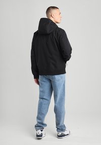 Black hooded jacket with a smooth texture, elastic cuffs, and a relaxed fit. Paired with light blue jeans and white-accented sneakers.