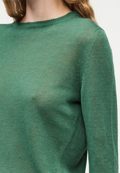 Gros plan d'une personne portant un pull à manches longues en maille fine vert avec un col rond sur un fond uni.