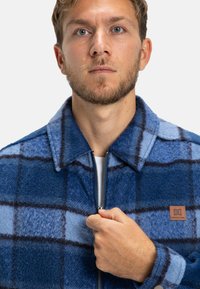 Blue and navy plaid jacket with a zip closure, made from textured fabric, featuring a collar and a branded leather patch on the chest.