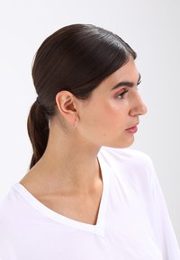Mulher jovem com cabelo escuro preso, usando um pequeno brinco de argola e uma camisa branca de decote em V, olhando para a direita sobre um fundo simples.