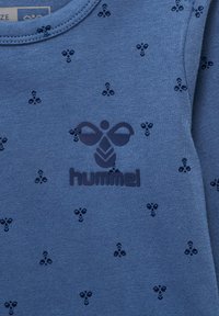 Niebieska bawełniana koszula z długim rękawem z powtarzającym się wzorem pszczół oraz ciemnoniebieskim logotypem "hummel" na środku. Gładka tekstura.