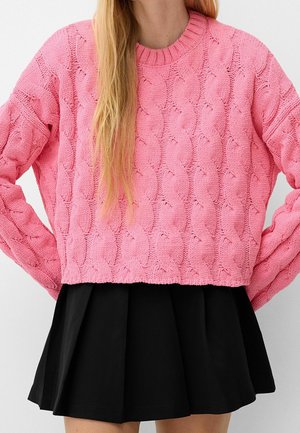 Pull en maille torsadée rose avec un col rond, coupe décontractée et poignets côtelés, associé à une jupe plissée noire. Détails de tissu texturé visibles.