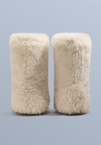 Puha krémszínű műszőrme lábszármelegítők, melyek bolyhos textúrájúak és egyszerű hengeres alakúak. Simított, díszítés nélküli kivitel.