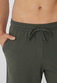 Olivově zelené jogger kalhoty mají strukturovanou tkaninu, elastický pas s šňůrkou a boční kapsy. Detailní záběr na pas a ruku v kapse.