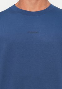 Close-up of a person wearing a blue crew neck shirt with small, subtle "Calvin Klein" logo embroidered on the chest.