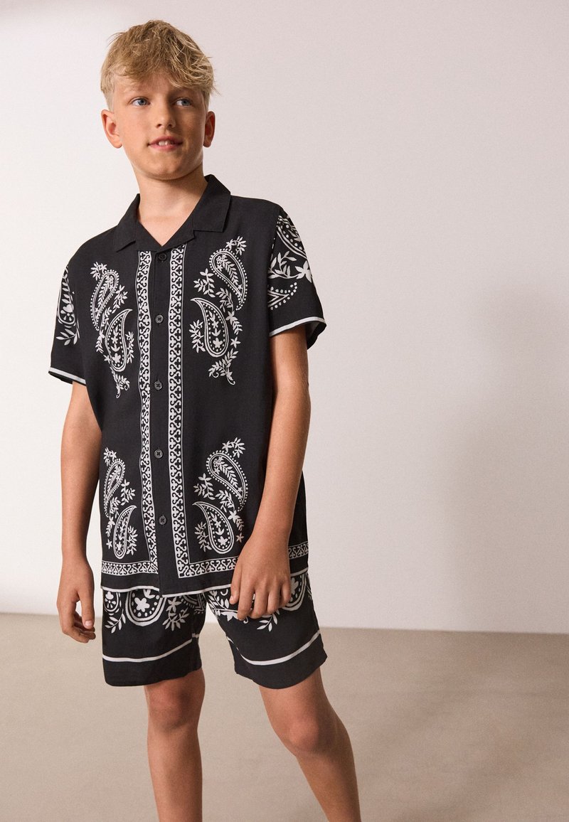Niño rubio con camisa negra de manga corta y pantalones cortos a juego con patrones blancos de paisley y flores, de pie contra un fondo claro y liso.