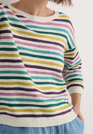 Femme portant un pull crème avec des rayures horizontales vertes, jaunes, violettes et roses, assorti à un jean bleu clair, main dans la poche.