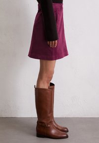 Jupe en velours côtelé violet au design droit, associée à des bottes en cuir marron hautes avec une sangle latérale et un détail de boucle.