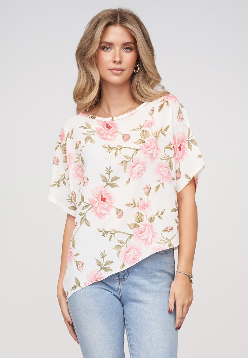 Femme aux cheveux blonds portant un chemisier blanc à manches courtes avec un imprimé floral rose et un jean bleu clair sur un fond uni.