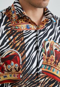 Chemise à manches courtes avec un motif de rayures de zèbre, ornée de couronnes royales, de détails ornés et de couleurs mélangées incluant le rouge, l'or et le noir.