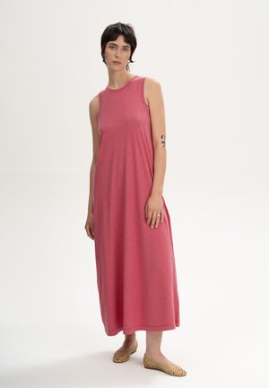 Jonge vrouw staand in een mouwloze roze jurk tot aan de enkels en beige gevlochten platte schoenen tegen een witte achtergrond.