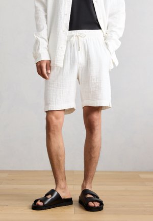 Uomo che indossa pantaloncini bianchi strutturati con coulisse, camicia bianca con bottoni, canotta nera e sandali neri aperti sulle dita su pavimento in legno.