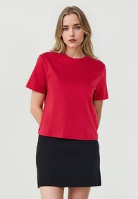 Jonge vrouw met blond haar draagt een rode top met korte mouwen en een zwarte rok, staand tegen een effen lichte achtergrond.