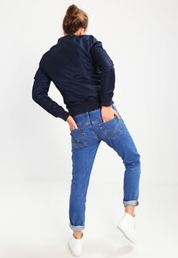 Chaqueta bomber marina con puños acanalados, combinada con jeans de mezclilla azul claro con dobladillos enrollados y bolsillos traseros. Zapatillas blancas completan el look.