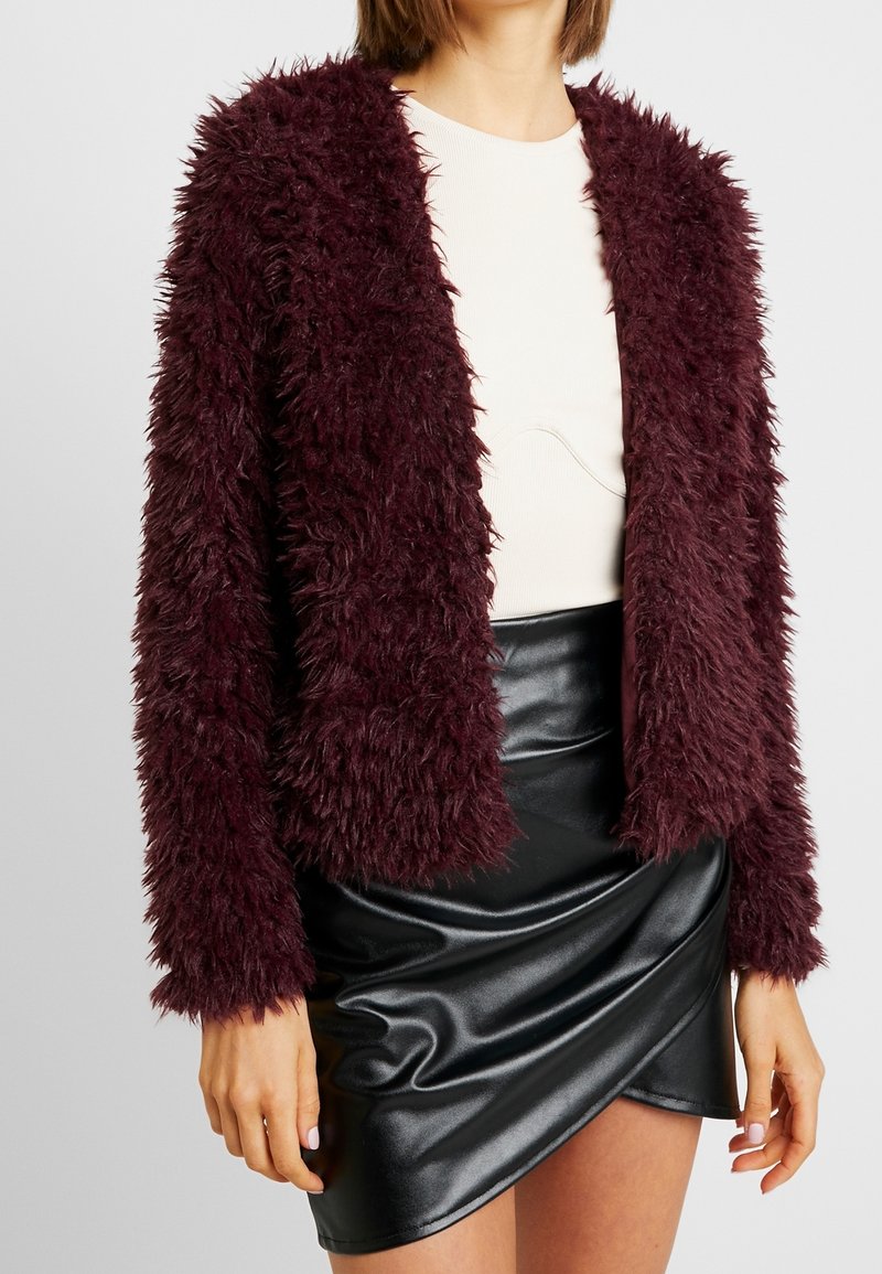 Burgundská umělá kožichová bunda s texturovaným povrchem, otevřeným designem vpředu a dlouhými rukávy, spárovaná s černou sukní z umělé kůže.
