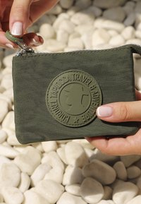 Pequeño estuche de color verde oliva con cremallera, hecho de tela texturizada, que presenta un destacado parche de goma redondo con un diseño en relieve.
