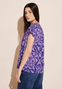 T-shirt viola a maniche corte con motivo astratto a contrasto e vestibilità comoda, abbinato a jeans blu scuro. Tessuto liscio, design informale.