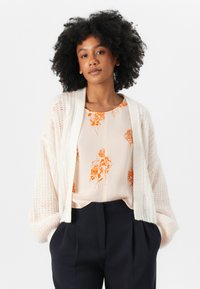Cardigan alb tricotat cu deschidere în față, purtat peste o bluză piersică deschisă cu modele florale portocalii, asociat cu pantaloni închiși la culoare.