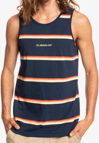 Homme portant un débardeur bleu marine avec des rayures horizontales orange et crème et le logo "Quiksilver" centré sur la poitrine. Mains dans les poches d'un short beige.