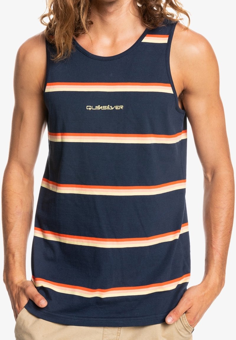 Homme portant un débardeur bleu marine avec des rayures horizontales orange et crème et le logo "Quiksilver" centré sur la poitrine. Mains dans les poches d'un short beige.