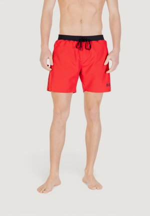 Man staat op blote voeten en draagt felrode zwembroek met zwarte elastische tailleband en trekkoord, tegen een effen lichte achtergrond.