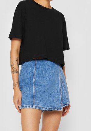 Kvinde iført sort kort T-shirt og kort blå denim nederdel, viser tatoveringer på venstre underarm, mod ensfarvet grå baggrund.